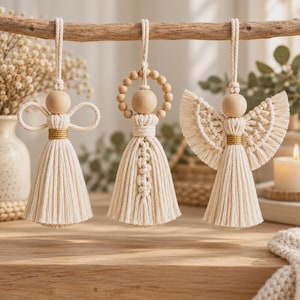May include: Three macrame angel ornaments hanging from a wooden branch. Each features a natural wooden bead head, off-white cotton cord body, and gold thread accents. The designs include looped wings, a beaded halo, and fringed wings.