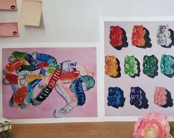 May include: Two framed prints of gummy candy art. The first print shows a pile of colourful gummy worms on a pink background. The second print shows a grid of nine gummy bears in different colours. The prints are signed by the artist, Angiemeart.