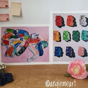 May include: Two framed prints of gummy candy art. The first print shows a pile of colorful gummy worms on a pink background. The second print shows a grid of nine gummy bears in different colors. The prints are signed by the artist, Angiemeart.