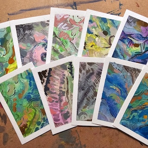 May include: Nine small, rectangular cards with abstract watercolor paintings in various colors, including blue, green, purple, and brown. The cards are arranged in a fan shape on a wooden surface.