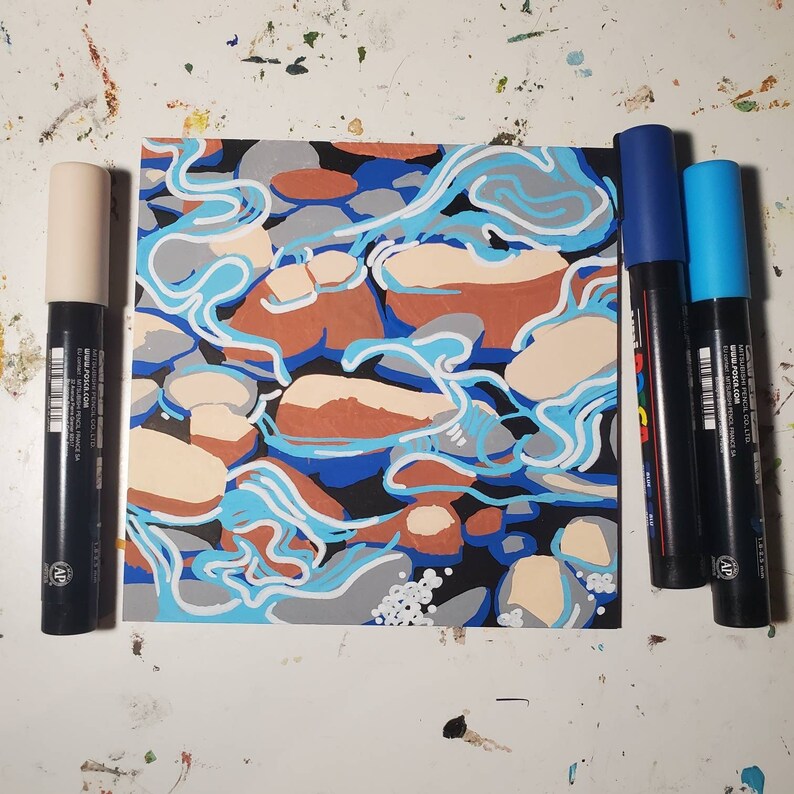 May include: A close-up of a small abstract painting with a blue, white, and brown color scheme. The painting depicts a swirling pattern of water and rocks. Three markers are visible in the background.