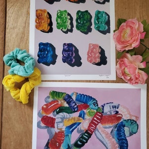 May include: Two prints of gummy candy paintings. The first print features a grid of 12 gummy bears in various colors, including red, orange, yellow, green, blue, and purple. The second print features a pile of gummy worms in various colors, including red, orange, yellow, green, blue, and purple. The prints are on a white background with a pink background.