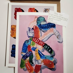 May include: A painting of a pile of colorful gummy worms on a pink background. The gummy worms are arranged in a random pattern and are of different shapes and sizes. The painting is signed "@angiemeart" in the bottom right corner.