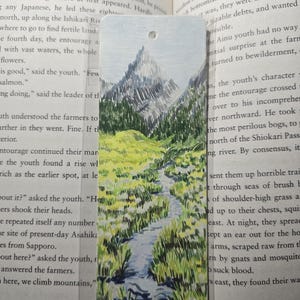 May include: A rectangular bookmark featuring a hand-drawn illustration of a mountain landscape. The scene depicts a tall, jagged mountain peak in the background with a winding blue stream flowing through a vibrant green grassy meadow in the foreground.