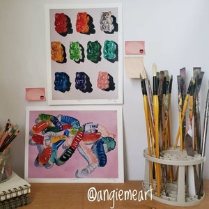 May include: Two framed paintings of colorful gummy candies, one with a pile of gummy worms and the other with a grid of gummy bears. The paintings are hanging on a white wall with a paintbrush holder in the foreground.