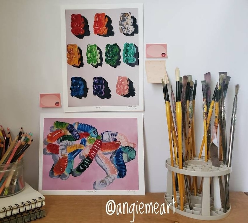 May include: Two framed paintings of colorful gummy candies, one with a pile of gummy worms and the other with a grid of gummy bears. The paintings are hanging on a white wall with a paintbrush holder in the foreground.