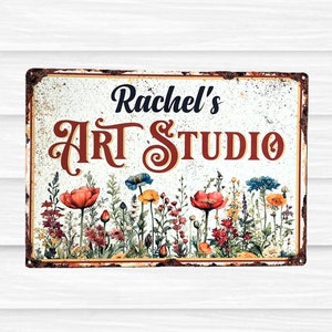May include: A rectangular metal sign with a distressed, rusted border featuring the text "Rachel's ART STUDIO" in black and dark red lettering. The lower portion displays a colourful illustration of various wildflowers in red, blue, and yellow.