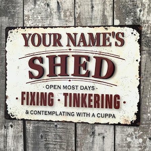 May include: White metal sign with red text that reads "YOUR NAME'S SHED - OPEN MOST DAYS - FIXING - TINKERING & CONTEMPLATING WITH A CUPPA".