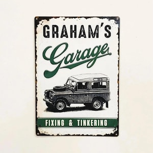 May include: A rectangular metal sign with a distressed white background featuring the black text GRAHAM'S and the green script text Garage. Below is a black and white illustration of a vintage off-road vehicle, with the text FIXING & TINKERING.
