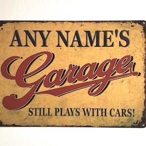 May include: A rectangular metal sign with a distressed yellow background and black and red text. The sign reads "ANY NAME'S Garage" in a vintage font, with "STILL PLAYS WITH CARS!" below.