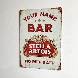 May include: A vintage-style metal sign with a white background and red text. The sign reads "Your Name Bar" with three stars above the word "Bar". Below the text is a Stella Artois logo with the text "Anno 1366 Stella Artois" and the words "No Riff Raff" at the bottom.
