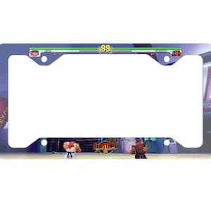 May include: A blue and white license plate frame featuring a video game scene from Street Fighter Alpha 3. The scene shows two characters fighting in a city setting. The text "Street Fighter Alpha 3" is visible in the center of the frame.