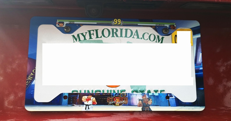 May include: A blue and white license plate frame with a green and white "MyFlorida.com" logo. The frame features a graphic of a video game character and the text "Sunshine State" and "3rd Strike: Fight for the Future".