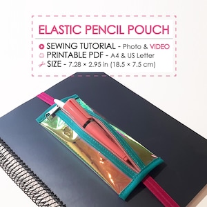 Puede incluir: Un estuche transparente e iridiscente con cremallera y ribete turquesa, que contiene un bolígrafo rosa. El estuche está sujeto a un cuaderno azul oscuro con una banda elástica rosa. El texto en la imagen dice "ELASTIC PENCIL POUCH" y "SIZE - 18.5 x 7.5 cm".