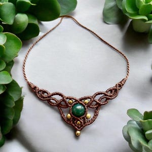 Peut inclure: Un collier macramé fait à la main avec une pierre verte centrale et des perles dorées. Le collier est fait de cordon marron et présente un design tissé complexe. Le collier est présenté sur une surface grise, avec des plantes vertes en arrière-plan.