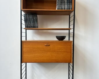 May include: A modular wall unit featuring a black metal ladder frame with wooden shelves and a central drop-down cabinet. The top shelf holds books, including one labeled PICASSO & CO., while a small black bowl sits on the cabinet surface.