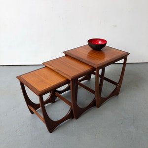 May include: A set of three nested wooden tables with square tops and curved, open-frame legs. The tables feature a warm brown wood grain finish. A small, round bowl with a matte black exterior and a glossy red interior sits on the largest table.