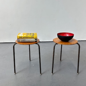 May include: Two round wooden stools with dark metal legs. The left stool holds two books titled Picasso & Co. and Holy Terror. The right stool holds a bowl with a matte black exterior and a vibrant red interior. The stools have a simple design.