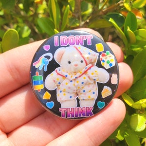 May include: A black and white button with a cartoon teddy bear wearing a polka dot outfit. The button says "I don't think" in pink and white letters.