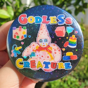 May include: A black button with a holographic finish featuring a pink and white clown with the text "Godless Creature" in rainbow colors. The button is surrounded by colorful toy blocks and other toys.