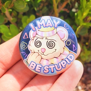 May include: A round button with a blue background and a white cartoon bear with a pink nose and pink and yellow striped collar. The bear has swirling eyes and is holding its paw to its mouth. The text "I MAY BE STUPID" is written in pink and yellow letters around the bear.