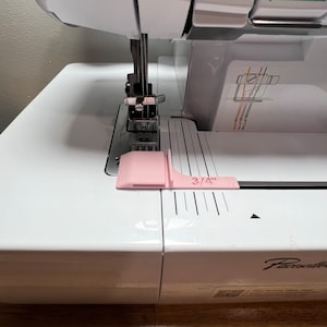 Puede incluir: Una máquina de coser blanca con una guía de costura rosa y una regla marcada en incrementos de 1/4 de pulgada. La máquina de coser tiene una aguja e hilo en su lugar, y la guía de costura está posicionada a la derecha de la aguja.