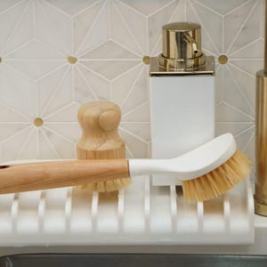 K&ouml;nnte beinhalten: Ein wei&szlig;er Sp&uuml;lb&uuml;rstenhalter mit einer B&uuml;rste mit Holzgriff, einem wei&szlig;en Seifenspender mit goldener Oberseite und einer Holzschrubbb&uuml;rste. Der Halter ist auf einer wei&szlig;en Arbeitsplatte mit einem goldenen Wasserhahn im Hintergrund montiert.