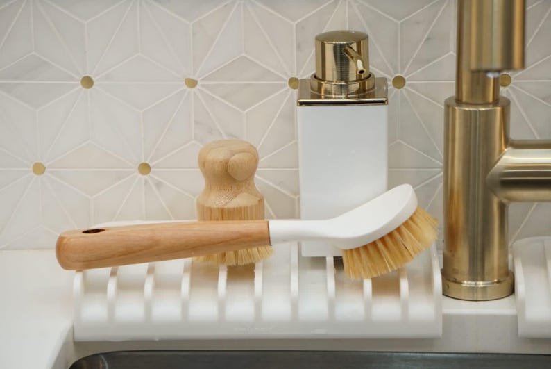 K&ouml;nnte beinhalten: Ein wei&szlig;er Sp&uuml;lb&uuml;rstenhalter mit einer B&uuml;rste mit Holzgriff, einem wei&szlig;en Seifenspender mit goldener Oberseite und einer Holzschrubbb&uuml;rste. Der Halter ist auf einer wei&szlig;en Arbeitsplatte mit einem goldenen Wasserhahn im Hintergrund montiert.