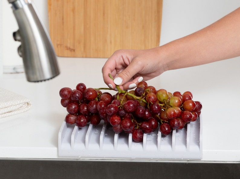 K&ouml;nnte beinhalten: Ein grauer Silikon-Obst- und Gem&uuml;seabtropfgestell mit roten Trauben darauf. Das Gestell hat mehrere Rippen, damit das Wasser abtropfen kann.