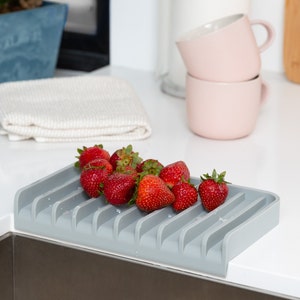 K&ouml;nnte beinhalten: Ein graues Silikon-Abtropfgestell mit abgerundeter Kante und mehreren Schlitzen zum Halten von Geschirr. Das Gestell ist mit frischen roten Erdbeeren gef&uuml;llt.