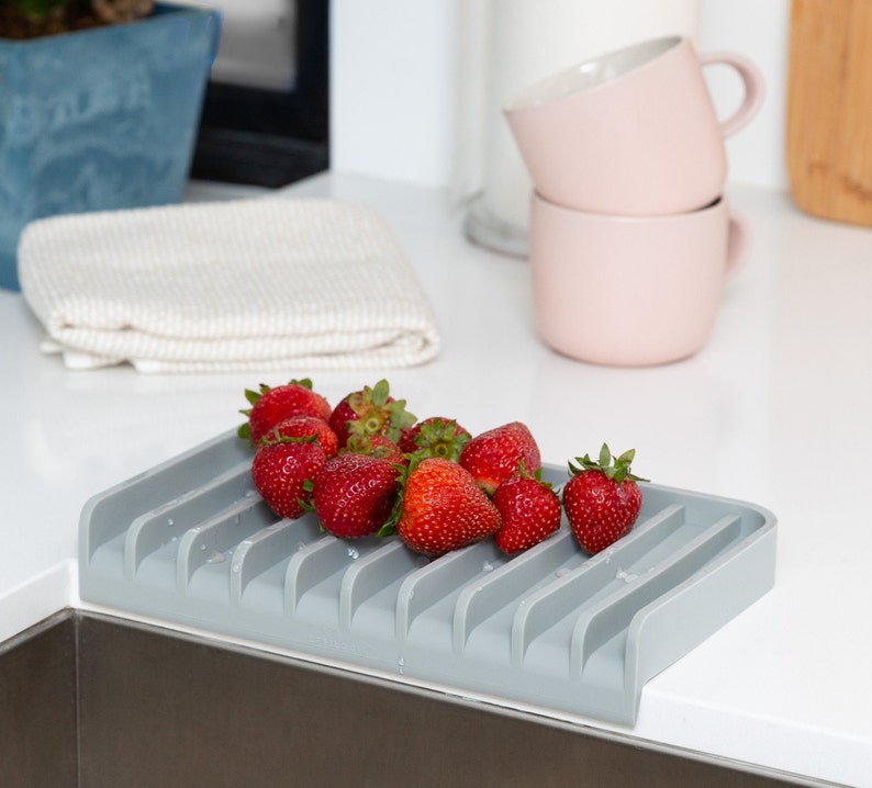 K&ouml;nnte beinhalten: Ein graues Silikon-Abtropfgestell mit abgerundeter Kante und mehreren Schlitzen zum Halten von Geschirr. Das Gestell ist mit frischen roten Erdbeeren gef&uuml;llt.