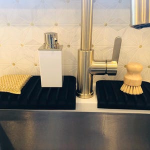 May include: Two black silicone dish drying racks with a gold faucet in the background. One rack has a yellow sponge and the other has a wooden brush.