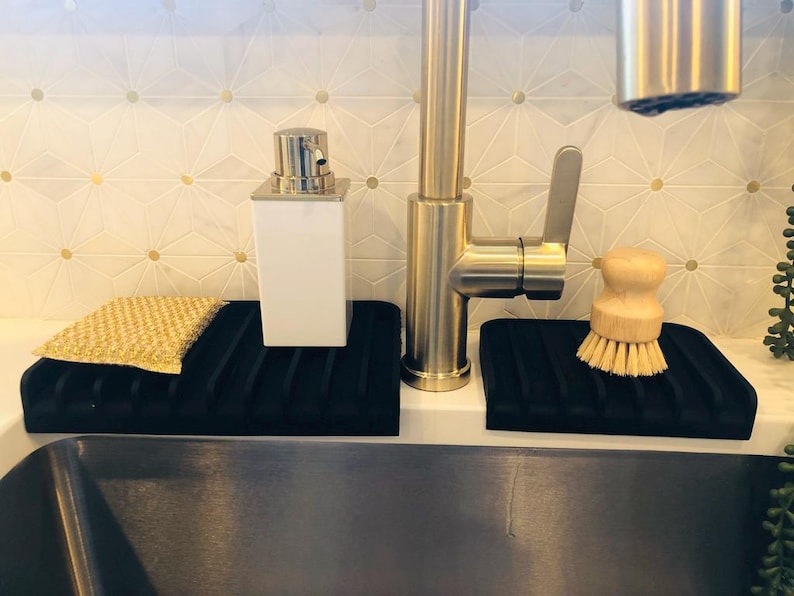 May include: Two black silicone dish drying racks with a gold faucet in the background. One rack has a yellow sponge and the other has a wooden brush.