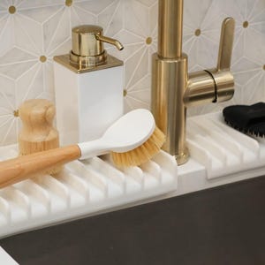 K&ouml;nnte beinhalten: Ein wei&szlig;er Abtropfst&auml;nder mit einer B&uuml;rste mit Holzgriff, einem wei&szlig;en Spender mit goldener Pumpe und einem schwarzen Schwamm. Der Abtropfst&auml;nder ist auf einer wei&szlig;en Arbeitsplatte montiert.