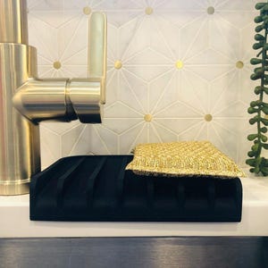May include: A black silicone dish drying rack with a gold sponge on top. The rack has multiple slots for drying dishes. The rack is sitting on a white countertop next to a gold faucet.