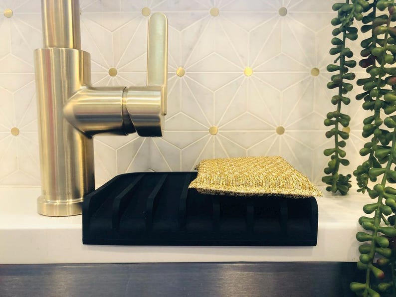May include: A black silicone dish drying rack with a gold sponge on top. The rack has multiple slots for drying dishes. The rack is sitting on a white countertop next to a gold faucet.