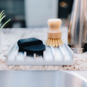 May include: A white plastic dish drying rack with a black sponge and a wooden brush. The rack is sitting on a granite countertop next to a kitchen sink.