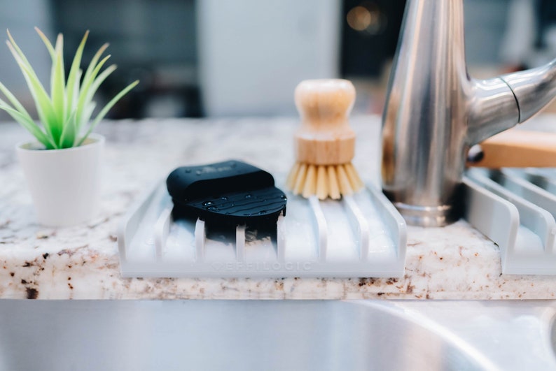 May include: A white plastic dish drying rack with a black sponge and a wooden brush. The rack is sitting on a granite countertop next to a kitchen sink.
