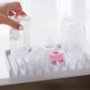 K&ouml;nnte beinhalten: Ein wei&szlig;er Babyflaschen-Abtropfst&auml;nder aus Kunststoff mit sechs durchsichtigen Glas-Babyflaschen und zwei durchsichtigen Plastik-Saugern. Eine Flasche tr&auml;gt das Etikett "Choice Organics".
