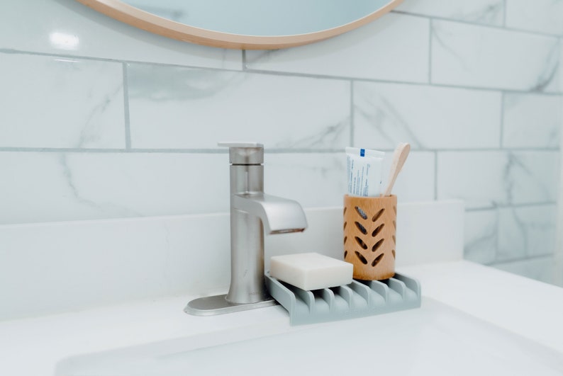 May include: A silver bathroom faucet with a white bar of soap and a wooden toothbrush holder with a tube of toothpaste. The faucet is mounted on a white countertop with white subway tile behind it.
