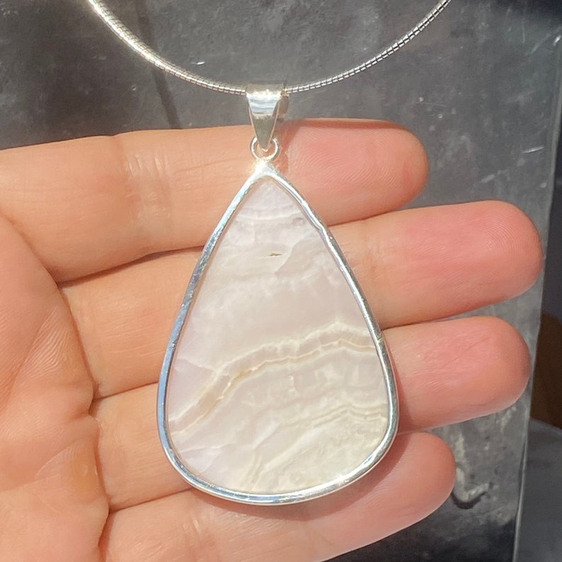 May include: A teardrop-shaped pendant with a white agate stone set in a silver frame. The pendant is hanging from a silver chain.