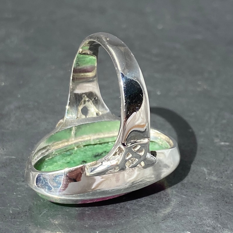 K&ouml;nnte beinhalten: Ein silberner Ring mit einem ovalen, gr&uuml;n-rosa Edelstein, der in einer silbernen Fassung sitzt. Der Ring hat ein detailliertes silbernes Band.