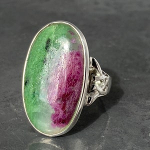 K&ouml;nnte beinhalten: Ein silberner Ring mit einem gro&szlig;en ovalen gr&uuml;n-rosa Edelstein. Der Ring hat ein keltisches Knotendesign an der Band.