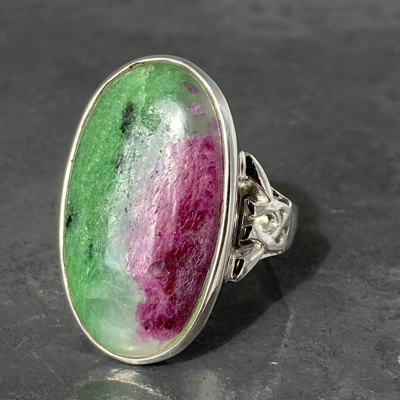 K&ouml;nnte beinhalten: Ein silberner Ring mit einem gro&szlig;en ovalen gr&uuml;n-rosa Edelstein. Der Ring hat ein keltisches Knotendesign an der Band.