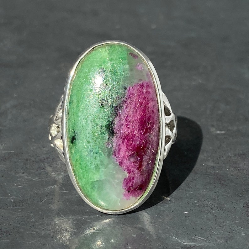 K&ouml;nnte beinhalten: Ein silberner Ring mit einem ovalen, gr&uuml;n-rosa Edelstein. Der Ring hat ein filigranes, kunstvolles Design.