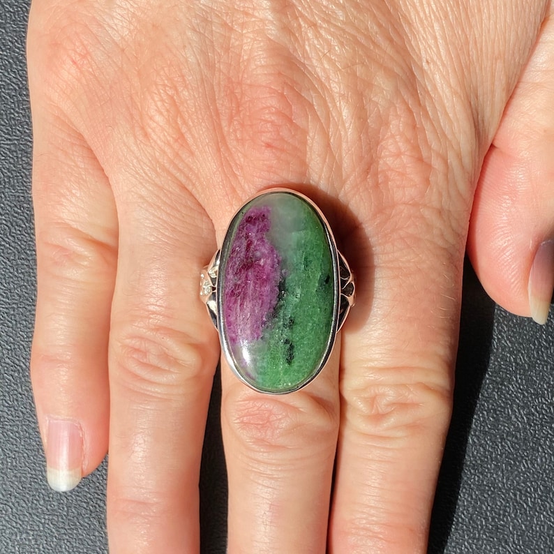 K&ouml;nnte beinhalten: Ein silberner Ring mit einem gro&szlig;en ovalen gr&uuml;nen und rosafarbenen Edelstein. Der Ring wird an einem Finger getragen.