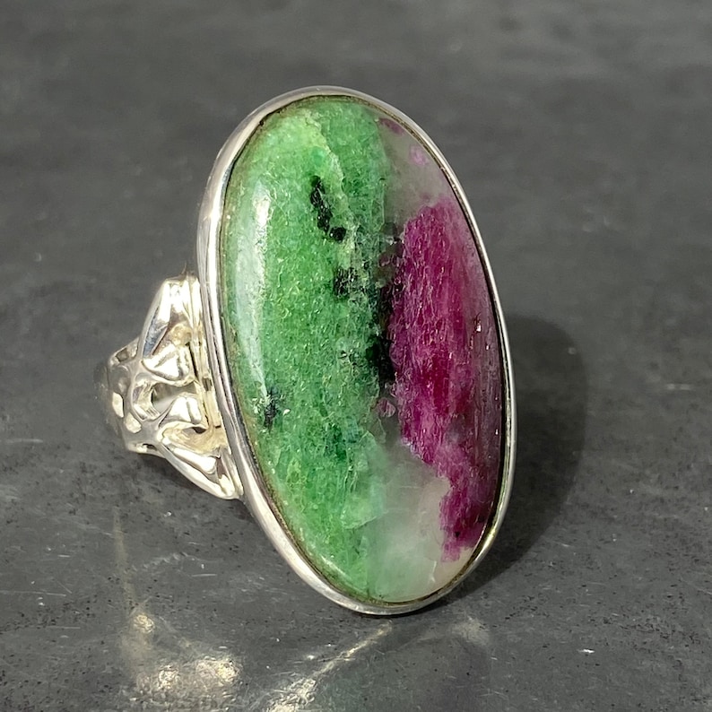 K&ouml;nnte beinhalten: Ein silberner Ring mit einem gro&szlig;en ovalen gr&uuml;n-rosa Edelstein. Der Edelstein hat ein geflecktes Muster aus Gr&uuml;n, Rosa und Wei&szlig;.