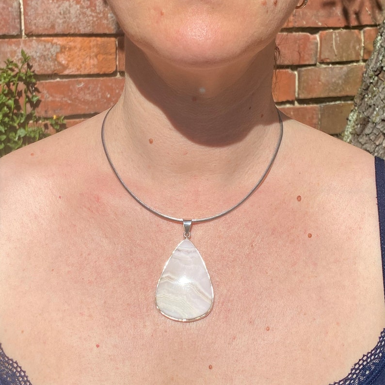 May include: A silver chain necklace with a large, teardrop-shaped pendant made of white agate. The pendant has a light pink and white swirl pattern.