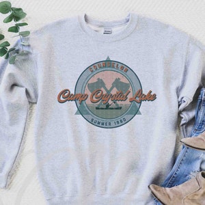 Op de afbeelding: Grijs sweatshirt met een vintage-stijl grafische afbeelding van een kamplogo. Het logo heeft een gekruist bijlontwerp, de tekst "Camp Crystal Lake" en de woorden "Counselor Summer 1980".