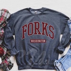 May include: A dark gray sweatshirt with the word "FORKS" in maroon letters with a white outline. Below the word "FORKS" is the word "WASHINGTON" in a smaller maroon font.